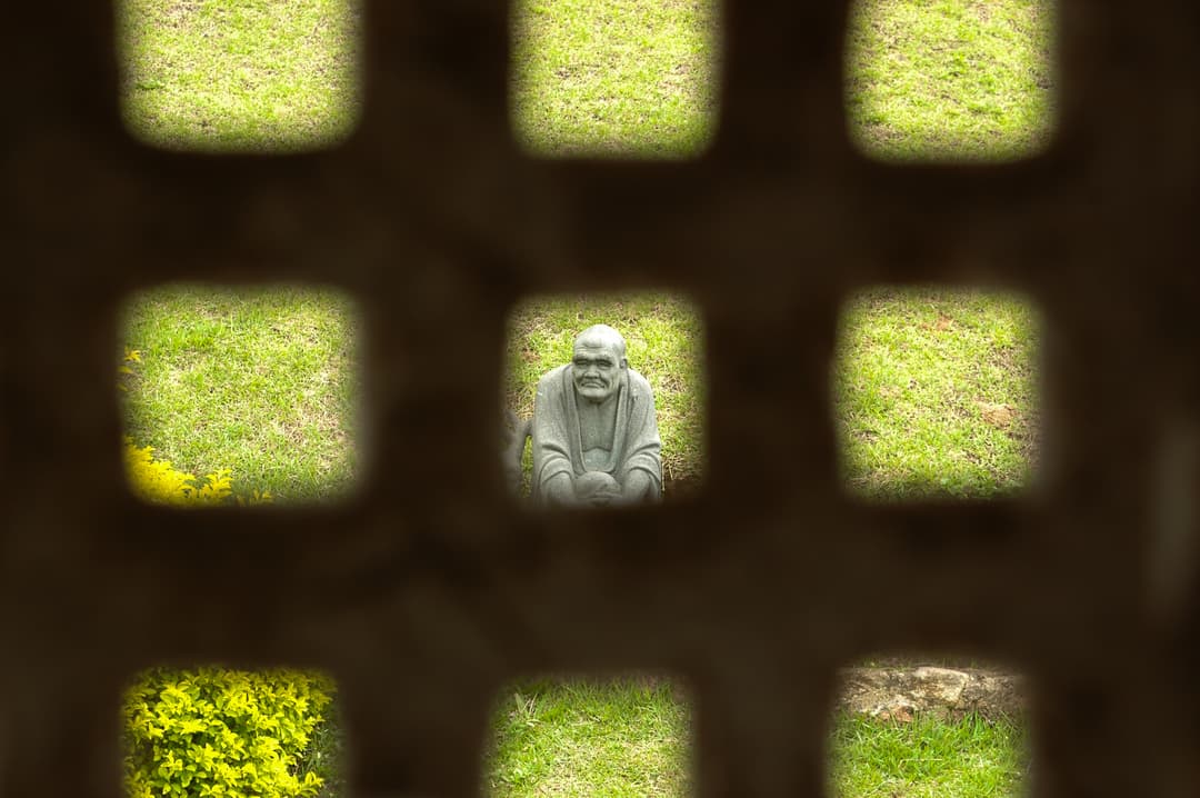 Stone monk statue viewed through a lattice screen in the temple garden.