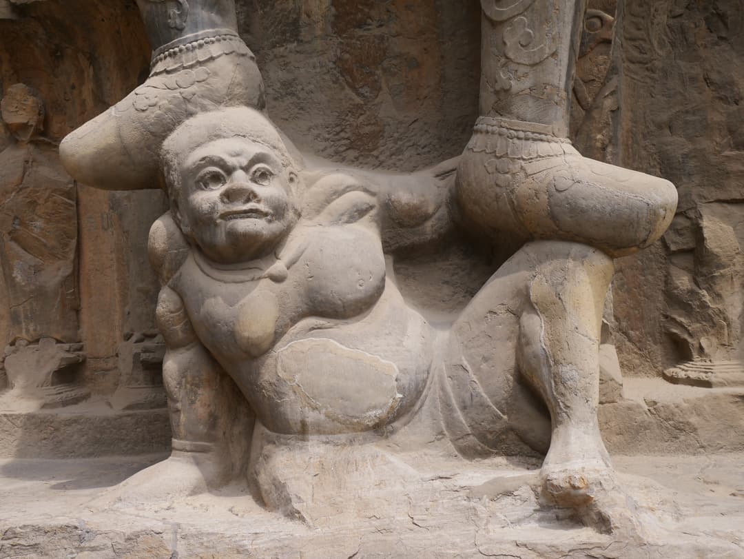 Detailed stone carving of a crouching yaksha supporting a guardian's feet.