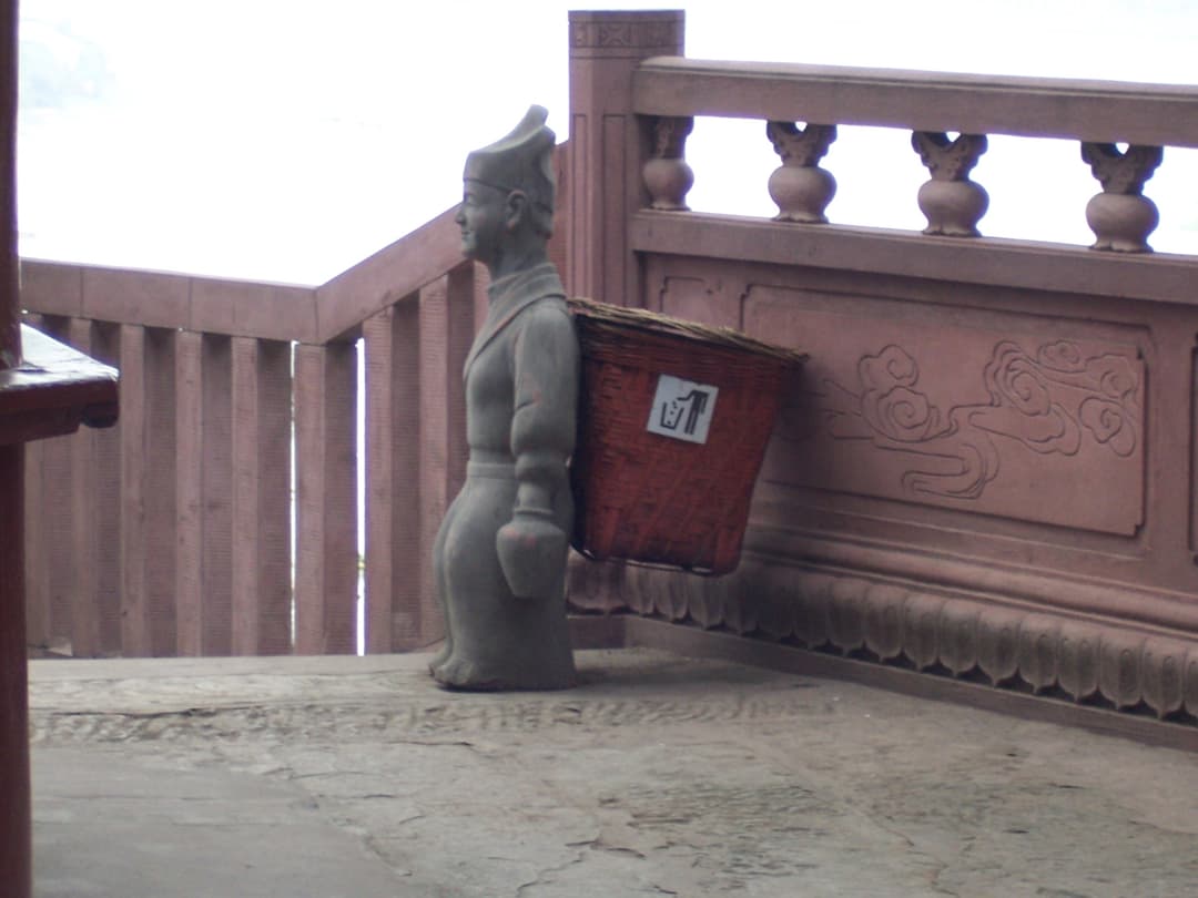 Statue-shaped trash bin with a back basket along a stone railing.