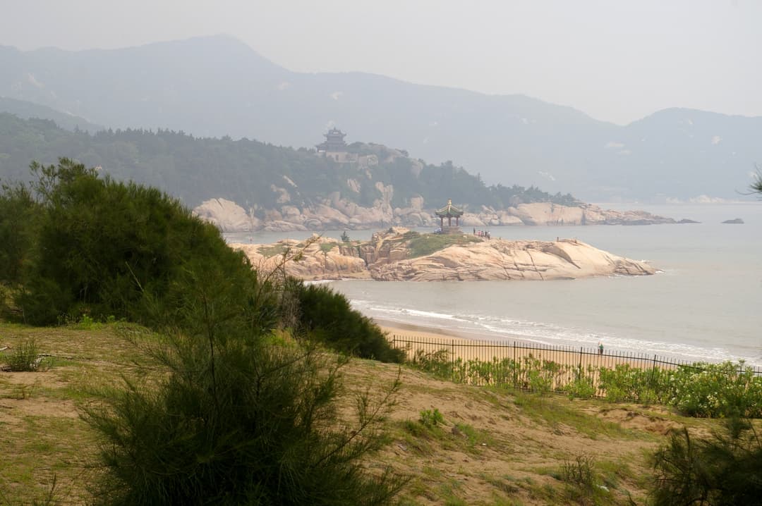 Wide view of a seaside pavilion on a rocky promontory below misty mountains.