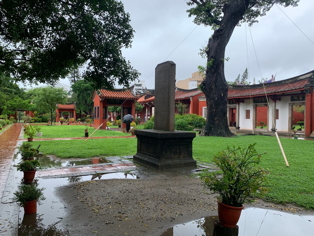 Courtyard grounds with a stone stele and traditional pavilions on a rainy day.