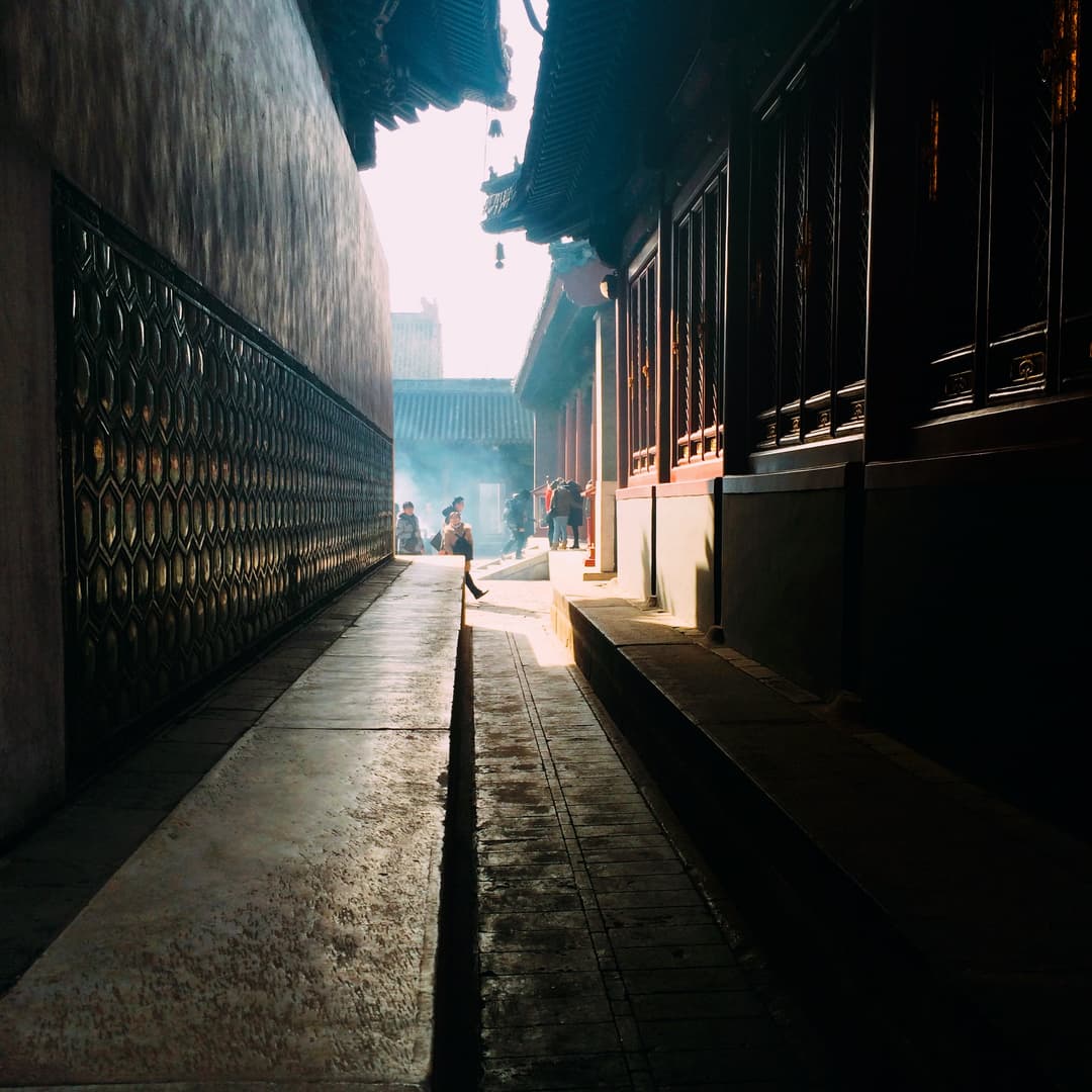 Shaded stone passageway leading into a sunlit, incense-filled courtyard.