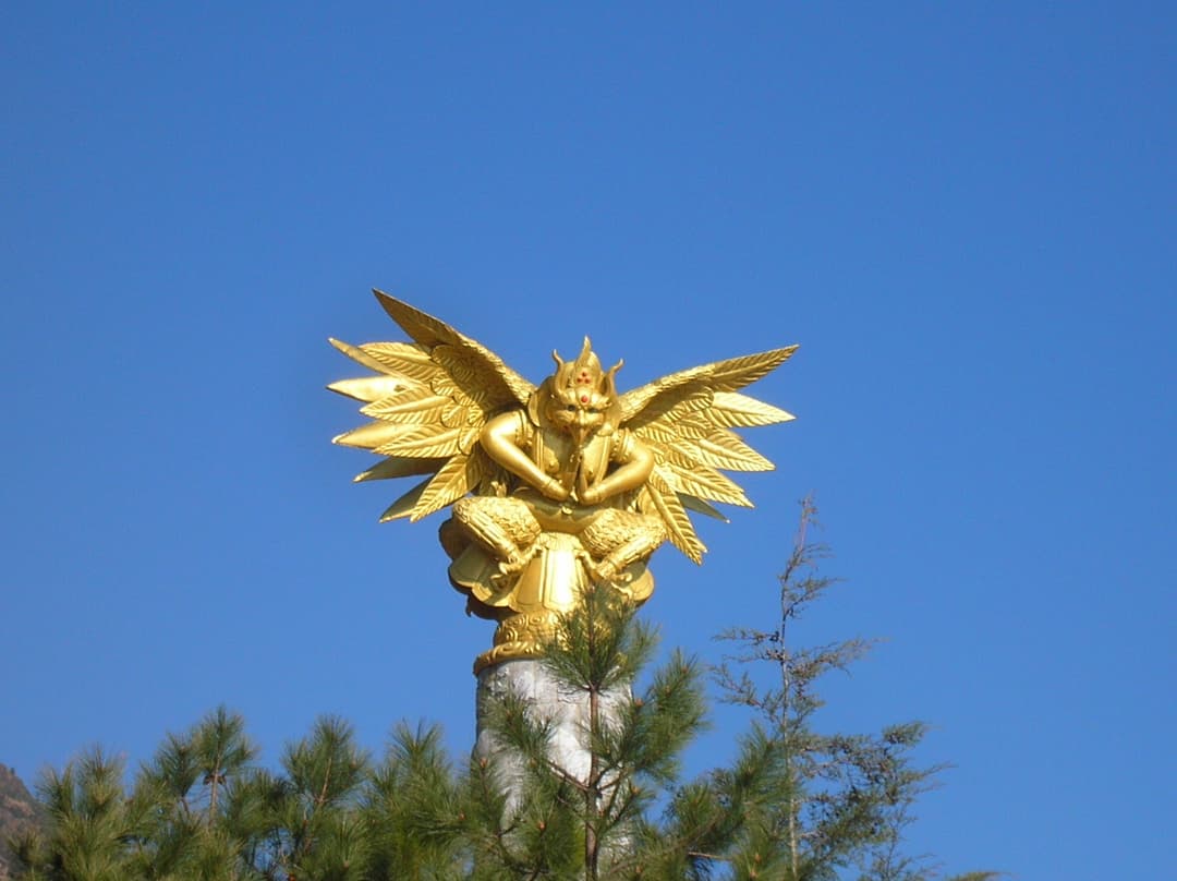 Golden Dongba Garuda statue atop a stone pillar against a clear blue sky.
