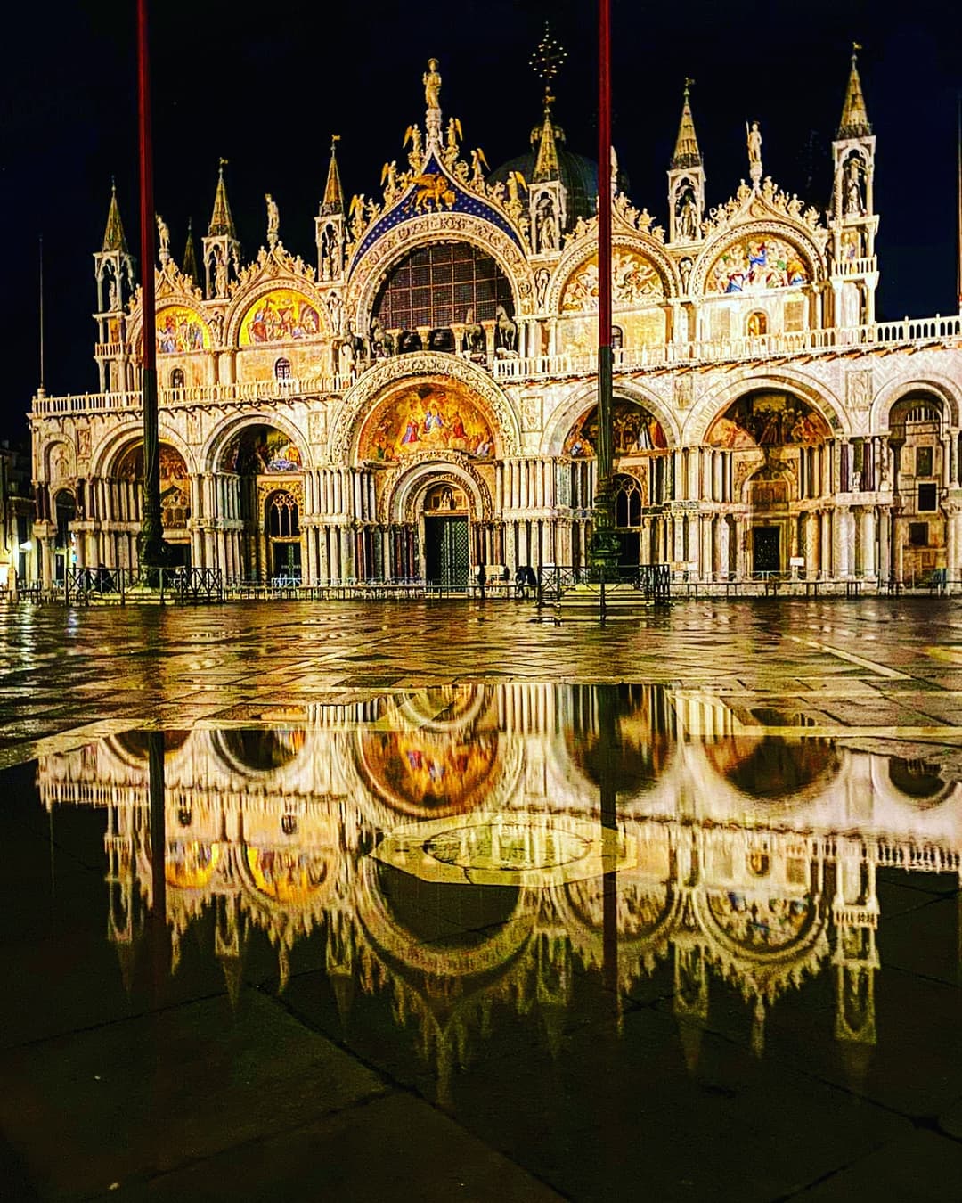Illuminated facade of St. Mark's Basilica reflected in water at night.