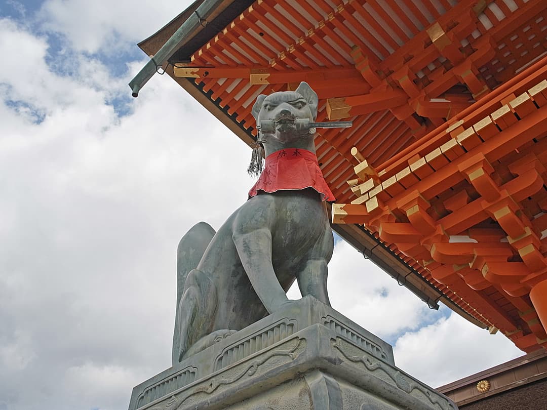 Low-angle view of a bronze kitsune statue under vermilion shrine eaves.