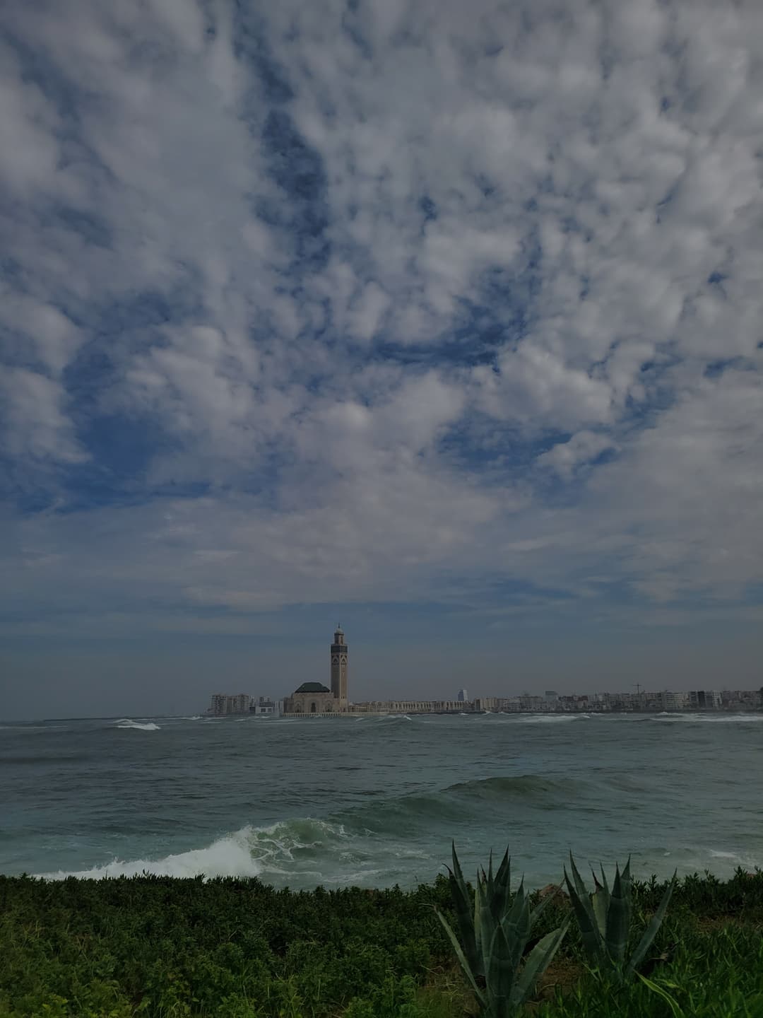 Distant view of Hassan II Mosque across Atlantic waves under a cloudy sky.