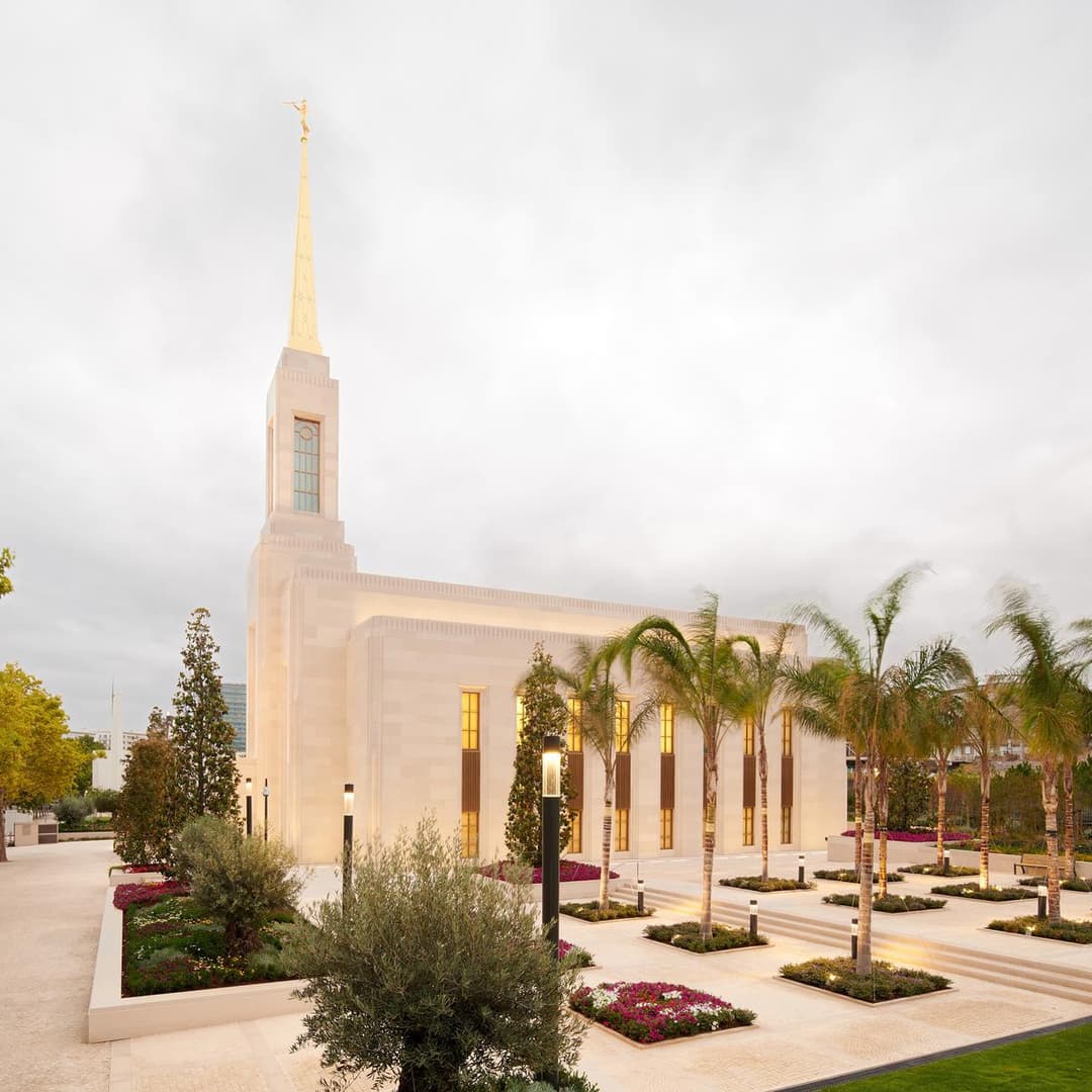 Twilight exterior of Lisbon Portugal Temple with spire, gardens, and palm trees.