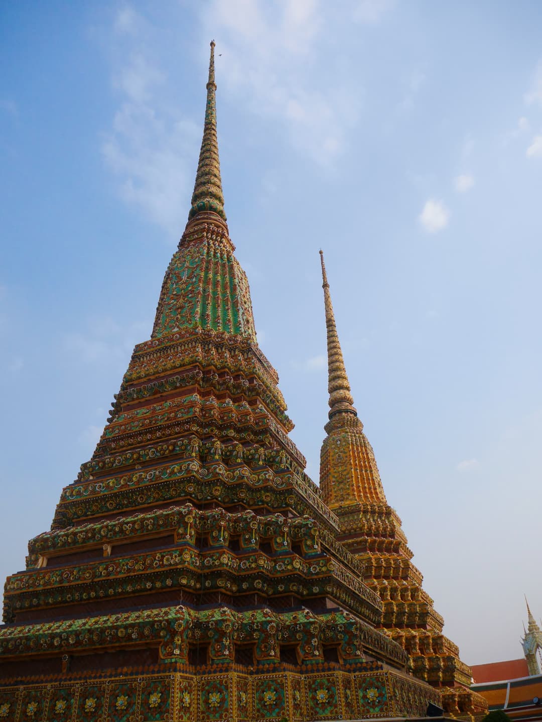 Low-angle view of two ornate mosaic-tiled chedis at Wat Pho against a blue sky.