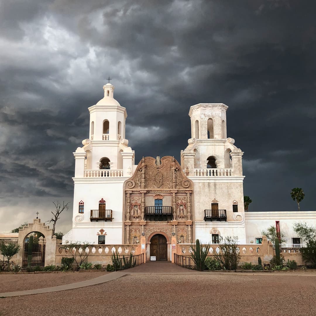 Front facade of Mission San Xavier del Bac under dramatic storm clouds.