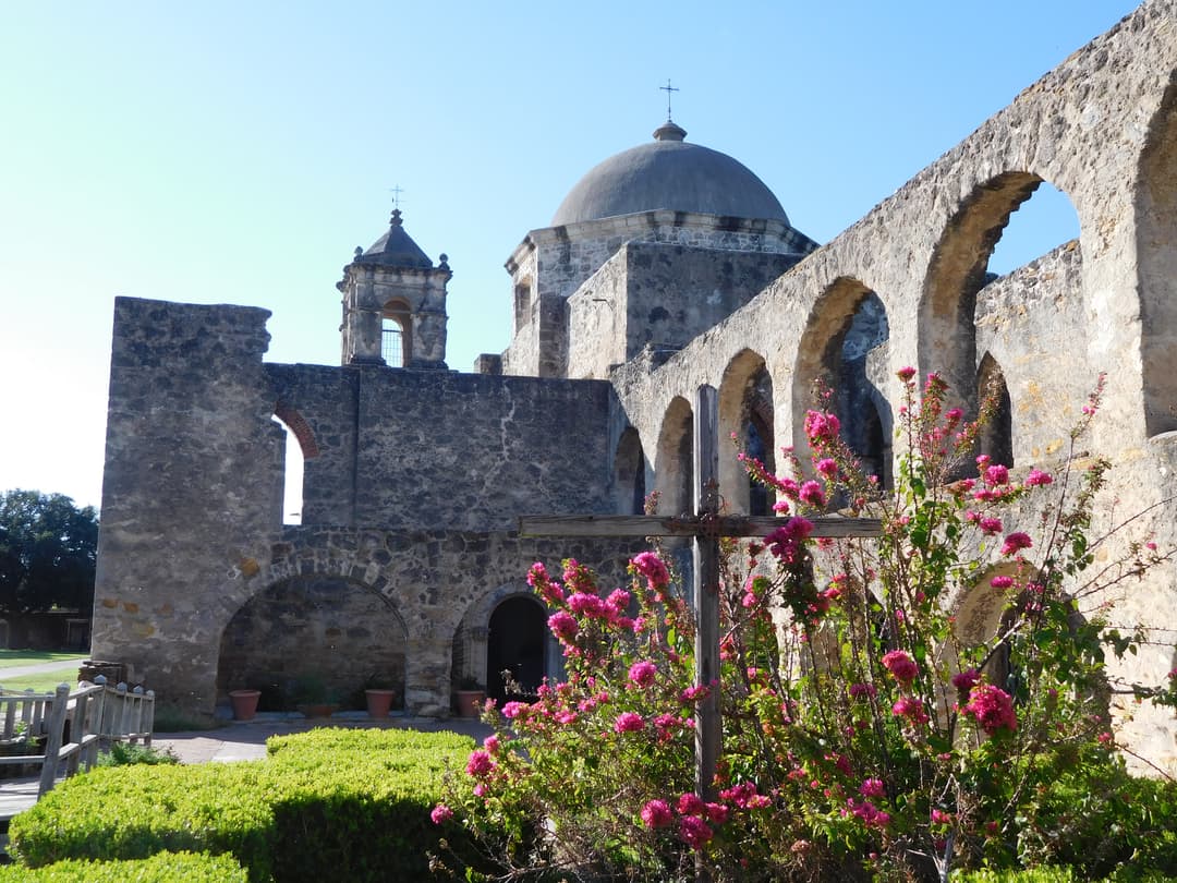 Stone arches, dome, and wooden cross framed by blooming pink flowers.