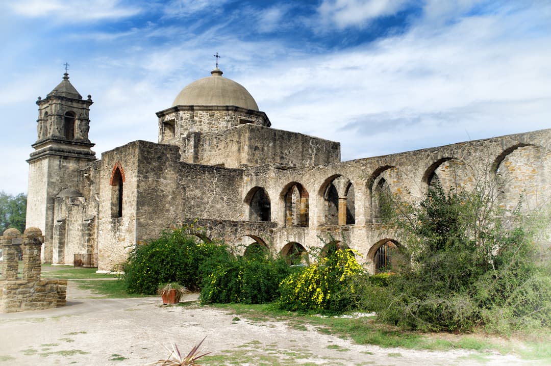 Wide exterior view of the stone mission, bell tower, dome, and arched cloister.