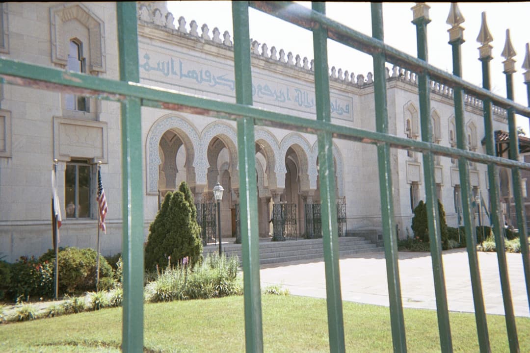 Front facade and arched portico framed by a green metal fence in daylight.