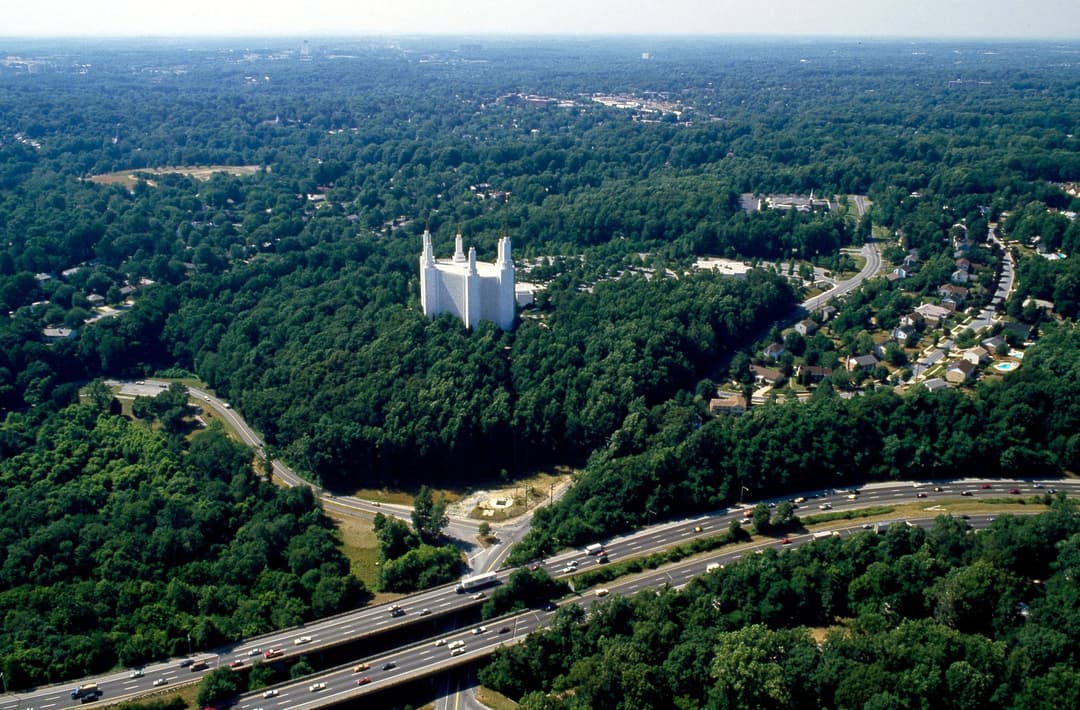 Aerial view of Washington D.C. Temple with six spires amidst lush trees and highways.