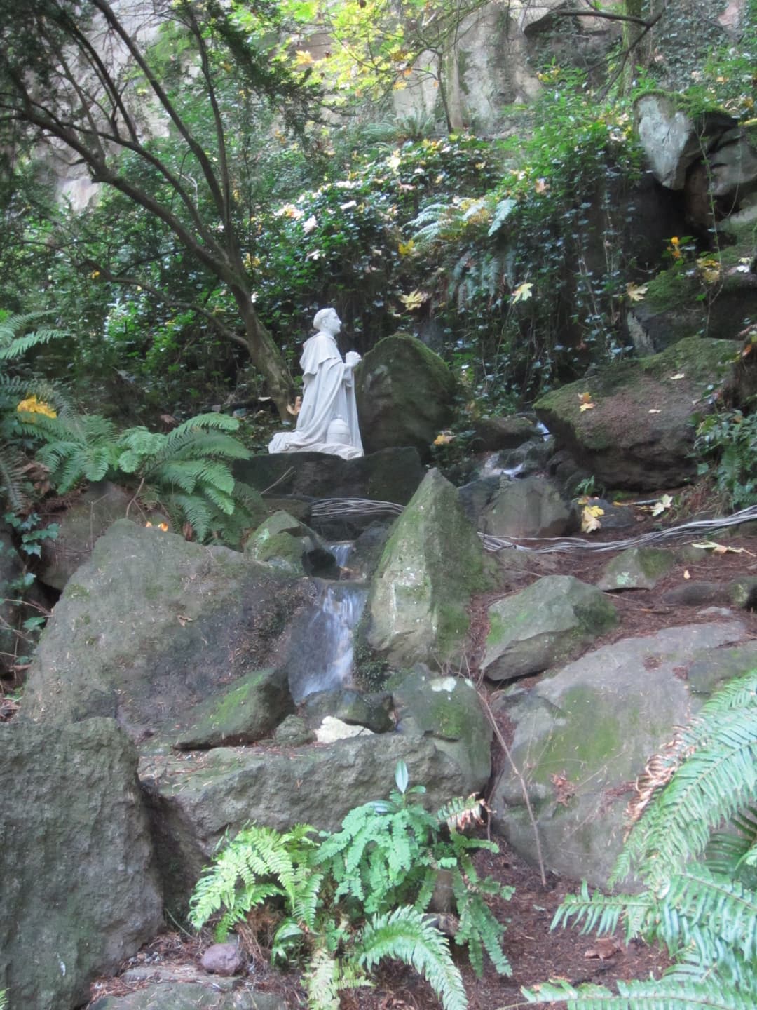 White statue kneeling in prayer beside a rocky waterfall amidst lush ferns.
