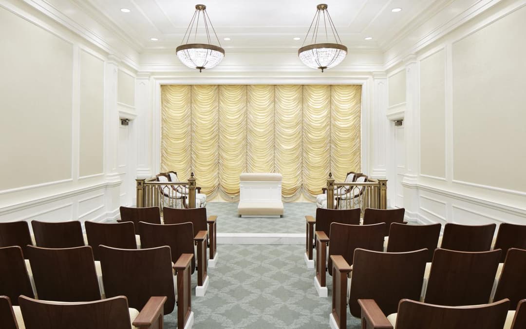 Interior of a temple room with rows of chairs, central altar, and chandeliers.