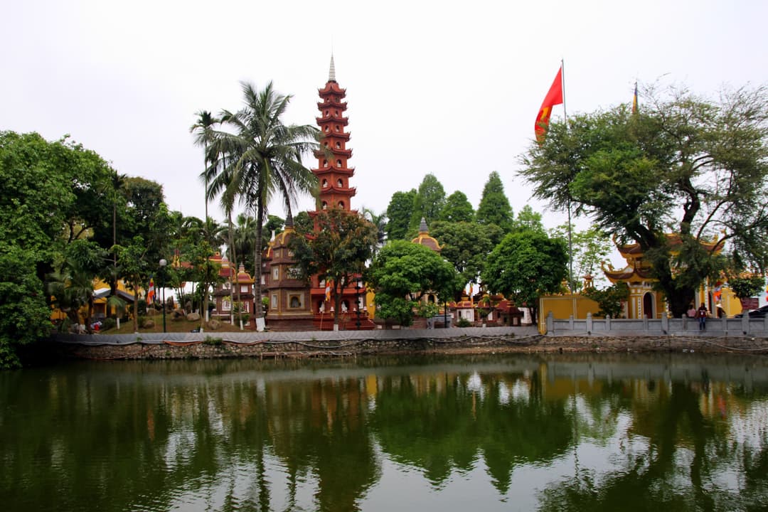 Wide exterior view of Trần Quốc Pagoda and red stupa reflected in the water.