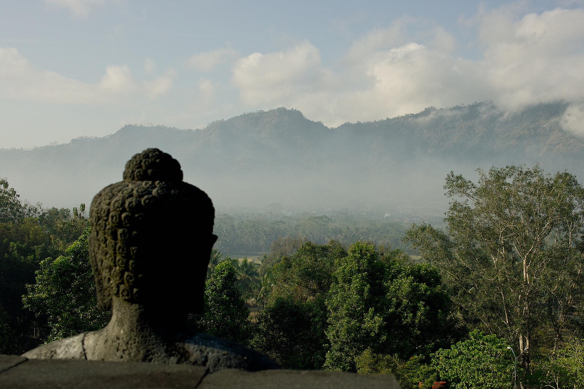A Buddha statue gazing over the misty Kedu Plain toward the Menoreh Hills at dawn.