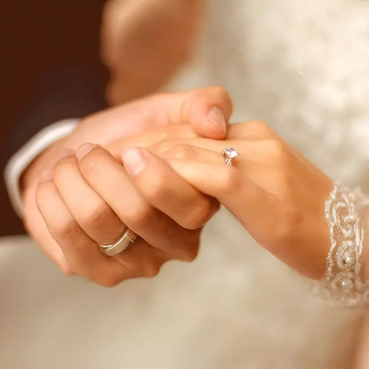 A man in a suit and a woman wearing a white dress holding hands, each with a ring on their finger.