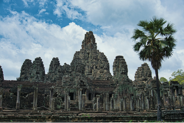 The Bayon Temple in Krong Siem Reap, Cambodia sits behind stone columns and a tall palm tree.