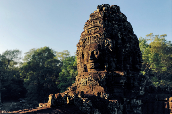 A deva, or diety, statue of a smiling face at Angkor Thom. Trees can be seen in the background beneath a pale blue sky.