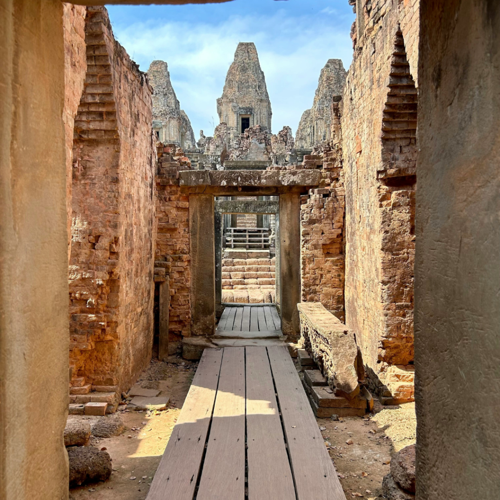 An outdoor corridor at the Angkor Wat Temple. Red brick archways surround the wooden plank path leadinging towards the main towers of the Angkor Wat Temple.