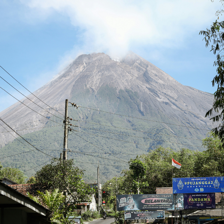 Mount Merapi in Central Java Indonesia sits in the distance, its peak seemingly touching the clouds. Power lines, a blue sign, and a street with a few cars can be seen in the foreground.