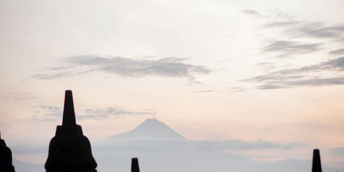 The stupa, or short temple spires of the Borobudur Temple sit in the foreground, while clouds, a misty blue mountain, and the pale orange sky can be seen in the background.