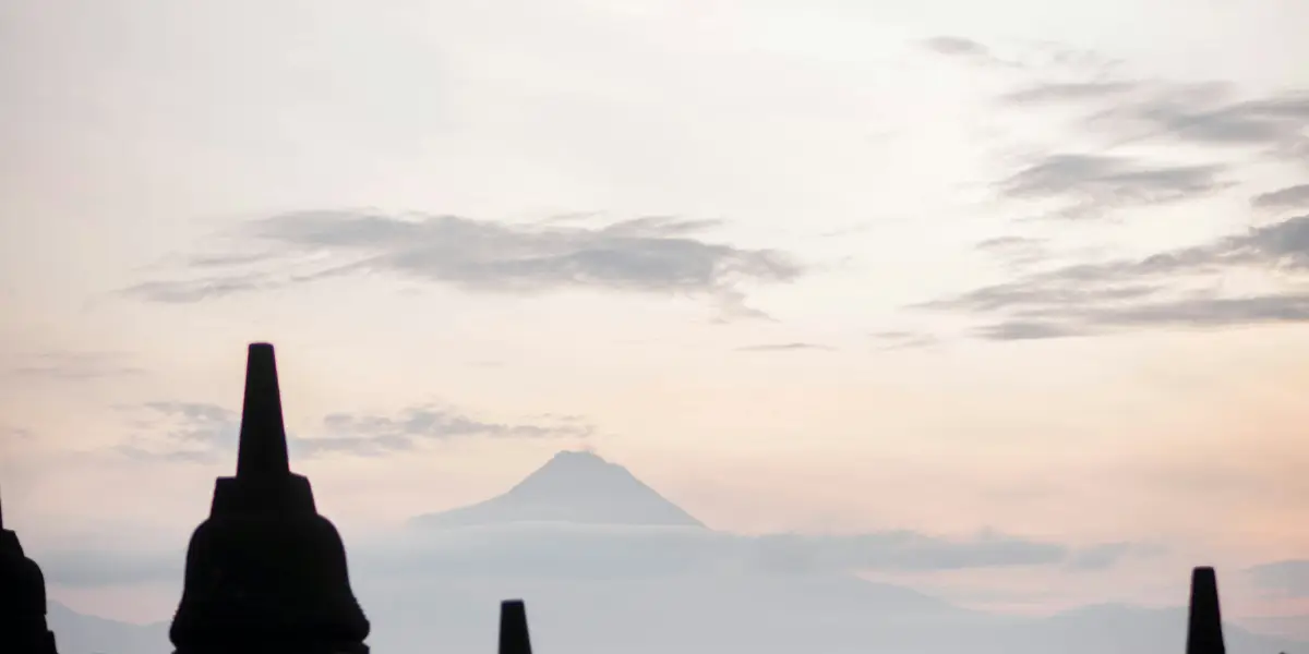 婆罗浮屠寺的佛塔或矮尖塔位于前景中，而背景中则可以看到云朵、雾蒙蒙的蓝色山脉和淡橙色的天空。