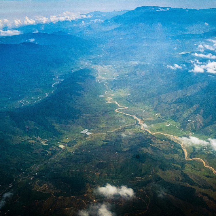 The dense green forests of Columbia are visible below. A river snakes its way through the lush, humid terrain below the pale blue sky and wisps of white clouds.