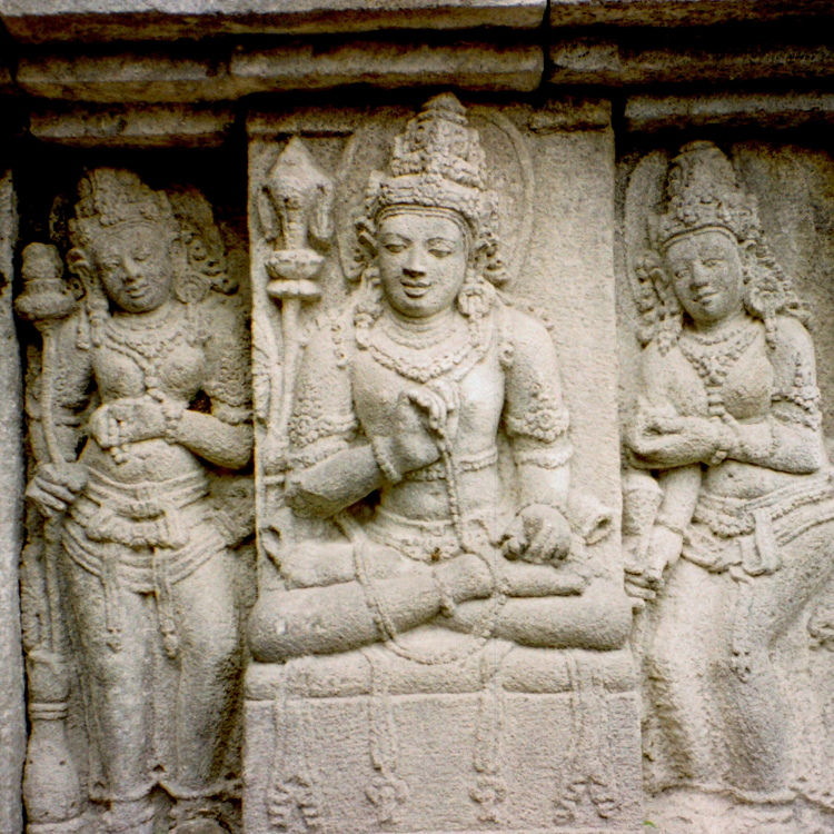 Three statues are engraved into stone at the Borobudur Temple, each wearing a crown. The middle statue sits with its legs crossed, while the other two statues stand on either side of the middle figure.