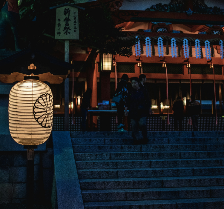 In the background, a group of onlooks sit or stand at the top of stone stairs outside a shrine at the Fushimi Inari Taisha Temple. j
