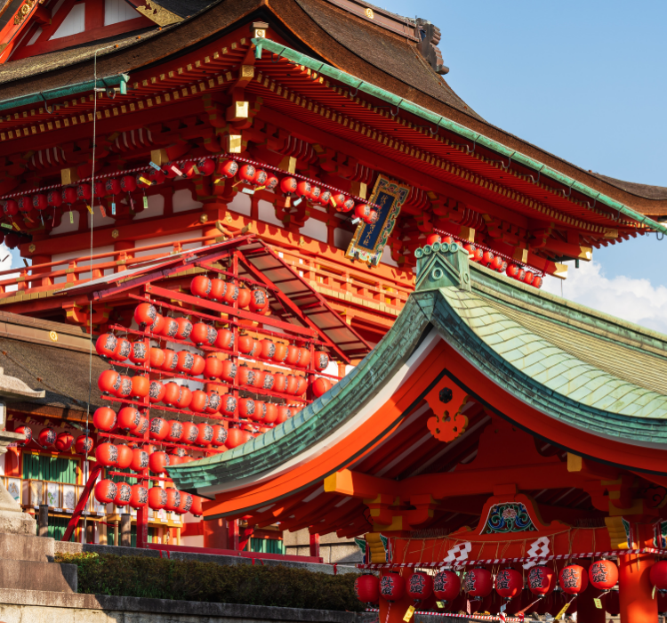 Two red buildings featuring curved, green-shackled roofs with traditional Japanese architecture. Several red paper lanterns hang from the two buildings and off another large red structure to the left at the Festival of Lanterns at the Fushimi Inari Taisha Shrine Temple.