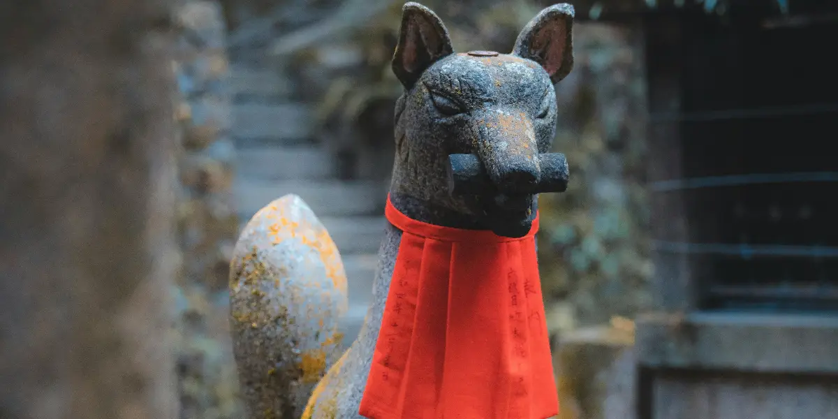 ثعلب حجري رمادي يرتدي وشاحًا أحمر وفي فمه مخطوطة إناري حجرية.