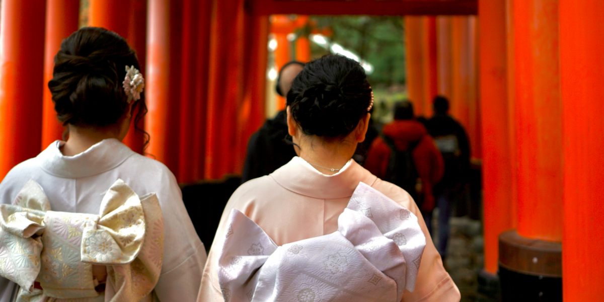 Two women wearing traditional ornate dresses strolling down a path beneath Torii gates. Each woman has a large bow made of intricate fabric on their back, their hair is styled with elaborate braids and buns.