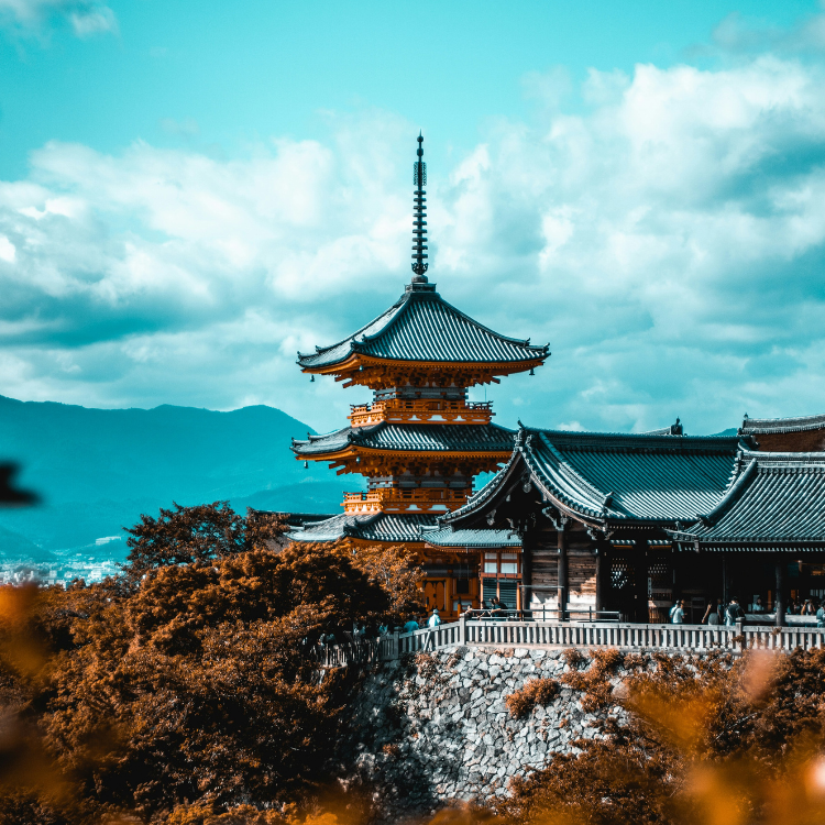 The Fushimi Inari Taisha Shrine with red and foliage in the foreground. A cyan-blue sky with fluffy clouds is visible in the background above the blue mountains.