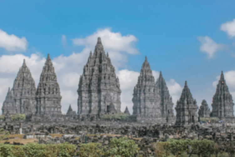 The Prambanan Temple​ can be seen, its gray spires pointing heavenward. Some bushes and grass are visible in the foreground.